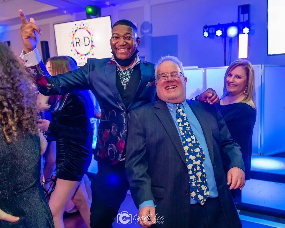 Man in a blue patterned jacket and older man in a suit and tie dancing with guests at a party