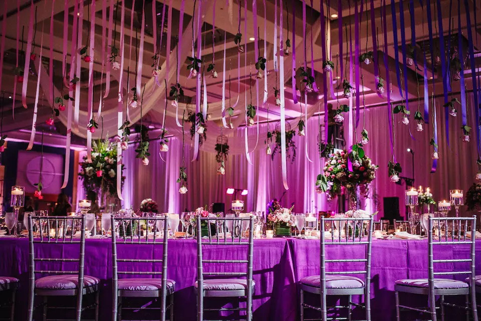 Ballroom with purple lighting, hanging ribbons and tables with silver chairs and floral centerpieces