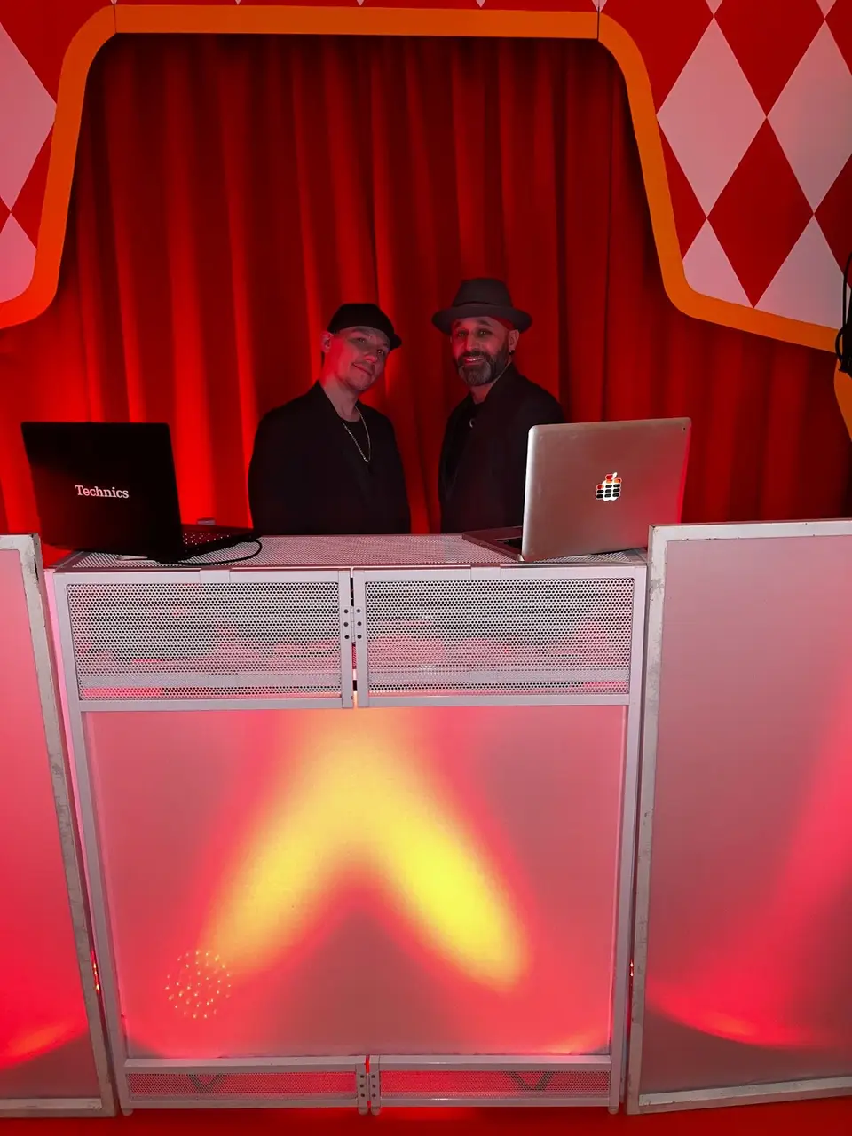 Two DJs in hats stand behind a DJ booth lit red and orange in front of a red curtain