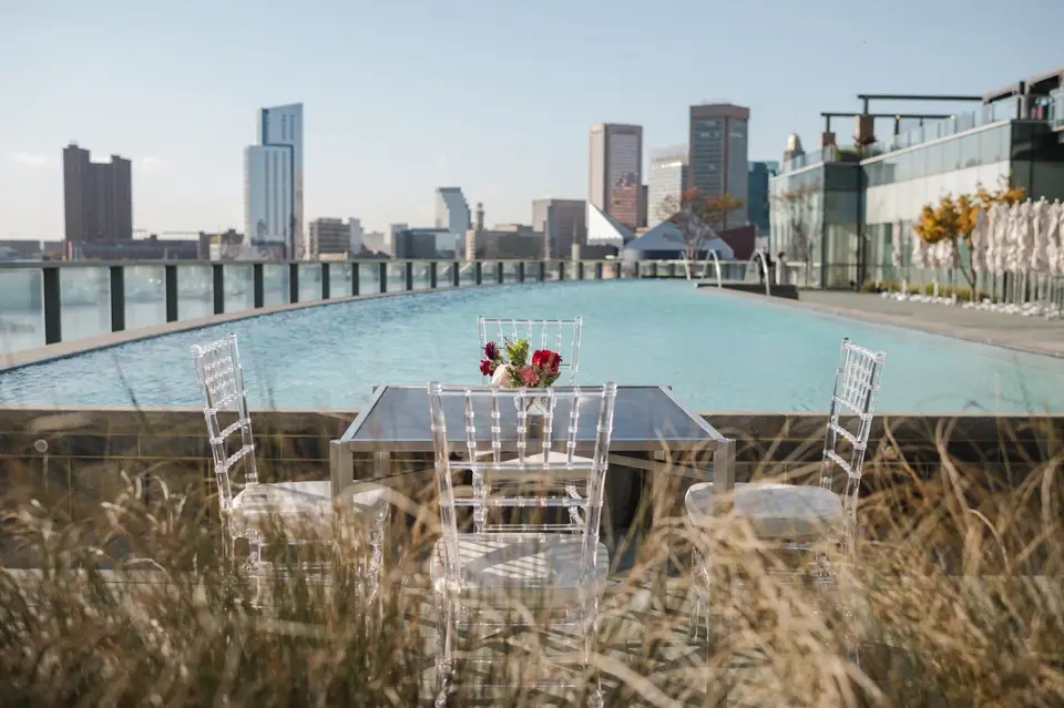 Clear chairs and a table with red flowers beside a rooftop pool with a city skyline behind