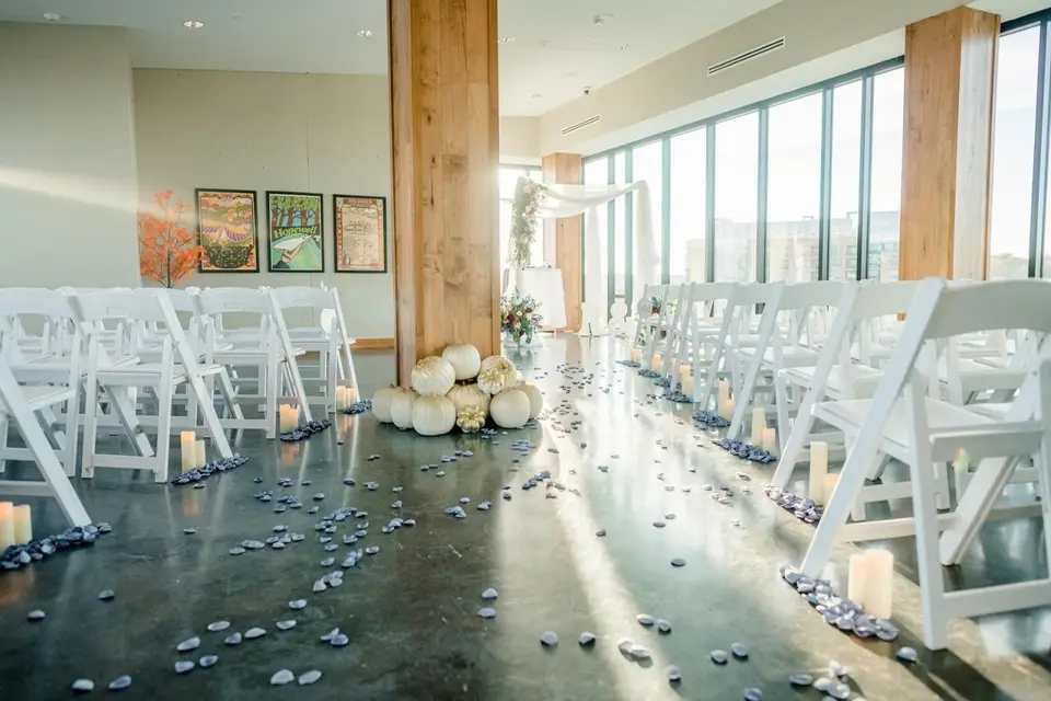 Wedding ceremony room with white chairs, pumpkins, candles and river stones along the aisle by large windows
