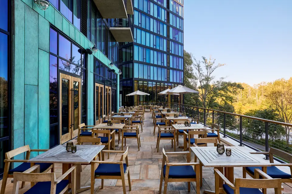 Outdoor hotel patio with wooden tables and chairs with blue cushions beside a glass building