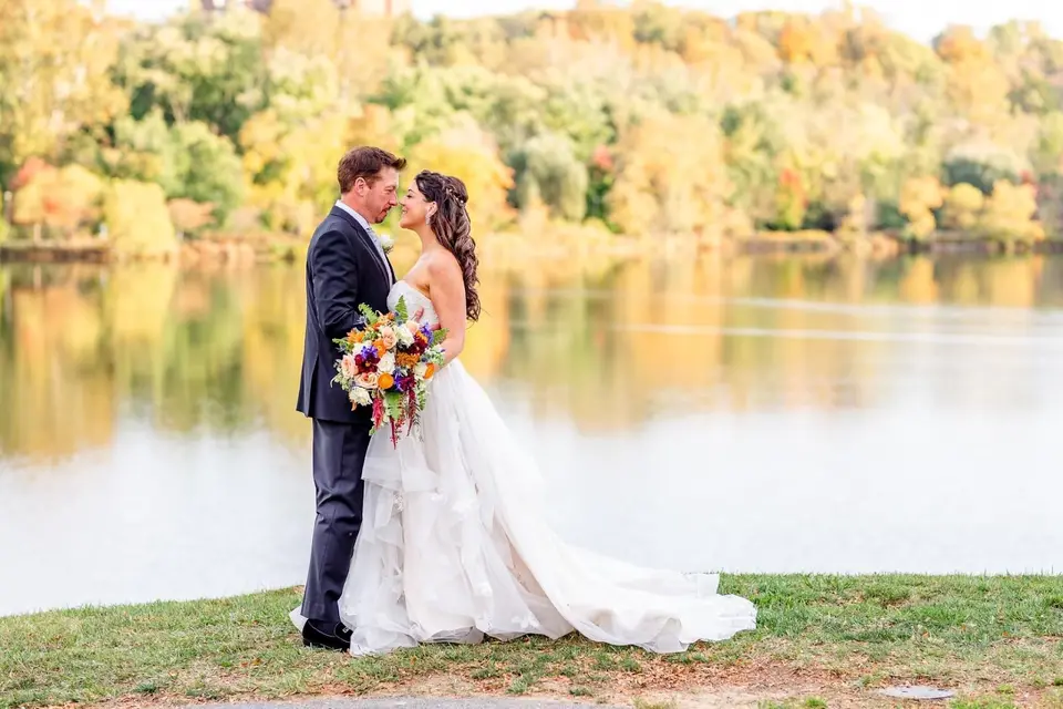 Bride and groom embracing on a grassy lakeshore with fall foliage across the water, bride holding a colorful bouquet
