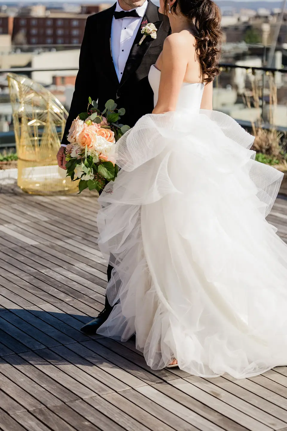 Groom in a tuxedo and bride in a layered tulle gown holding peach flowers on a wooden rooftop deck
