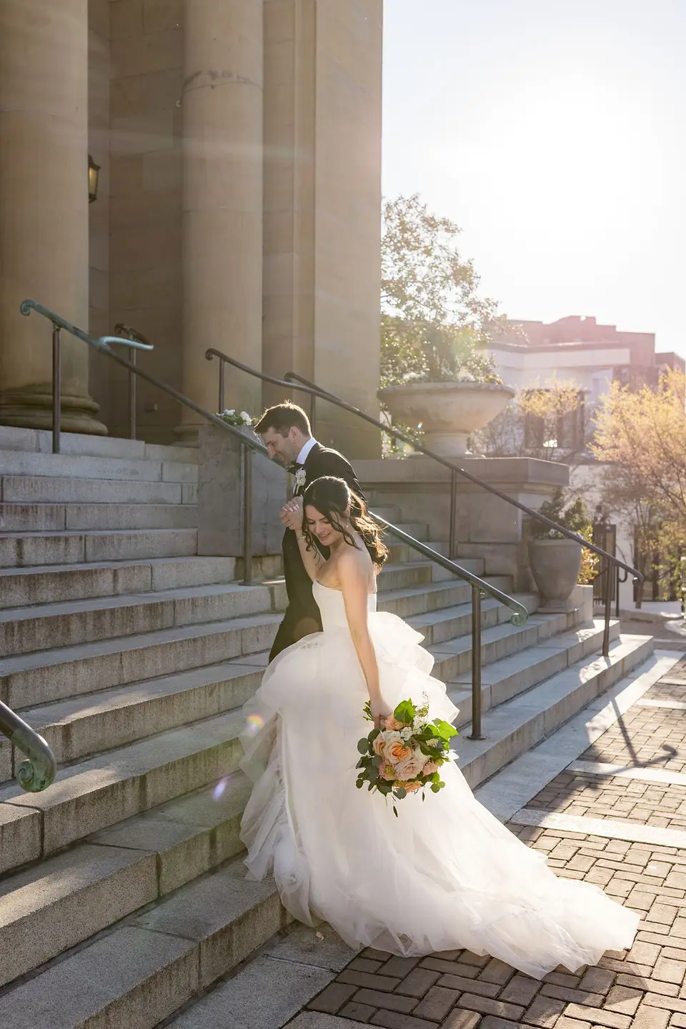 Bride and groom walk down stone steps by tall columns in the sunlight, bride holding a bouquet