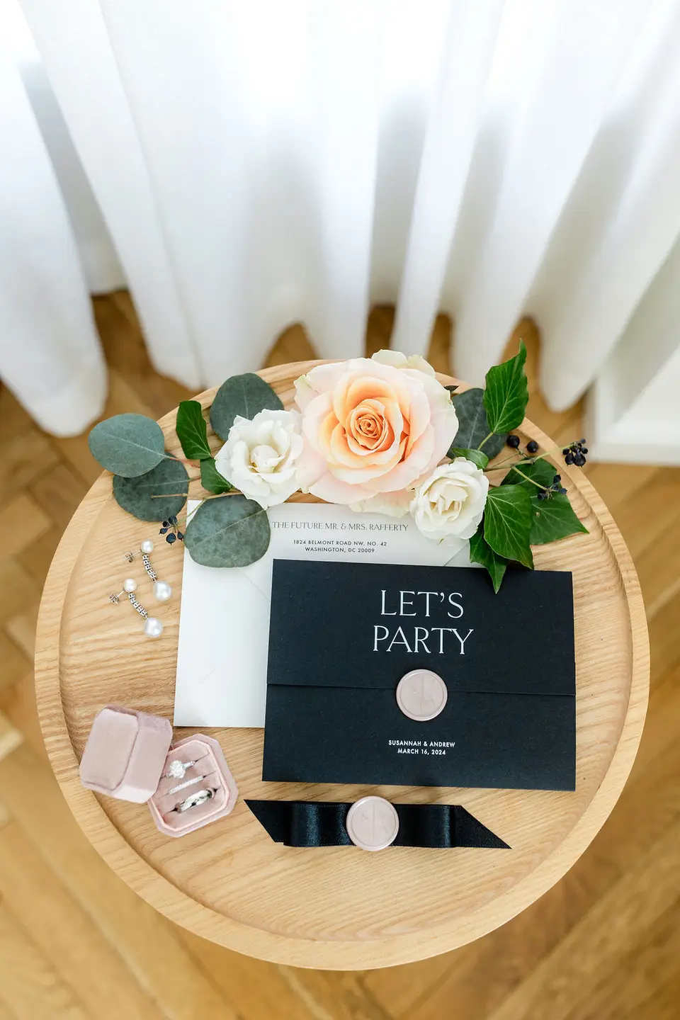 Black Let's Party wedding invitation with a wax seal, flowers, and a ring box on a round table