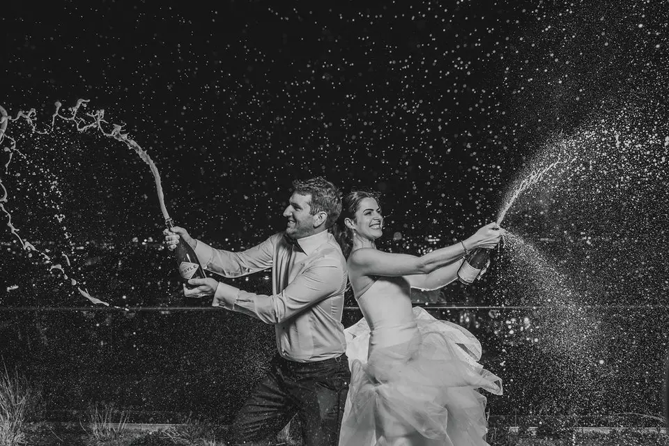 Black and white shot of a bride and groom back to back spraying champagne at night