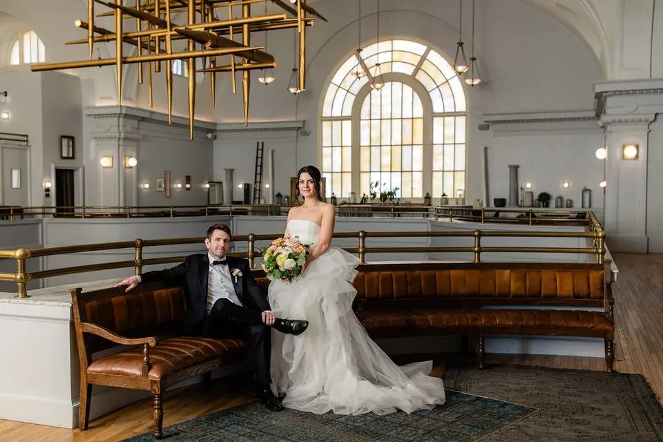 Bride sitting and groom reclining on leather benches in a bright hall with arched windows