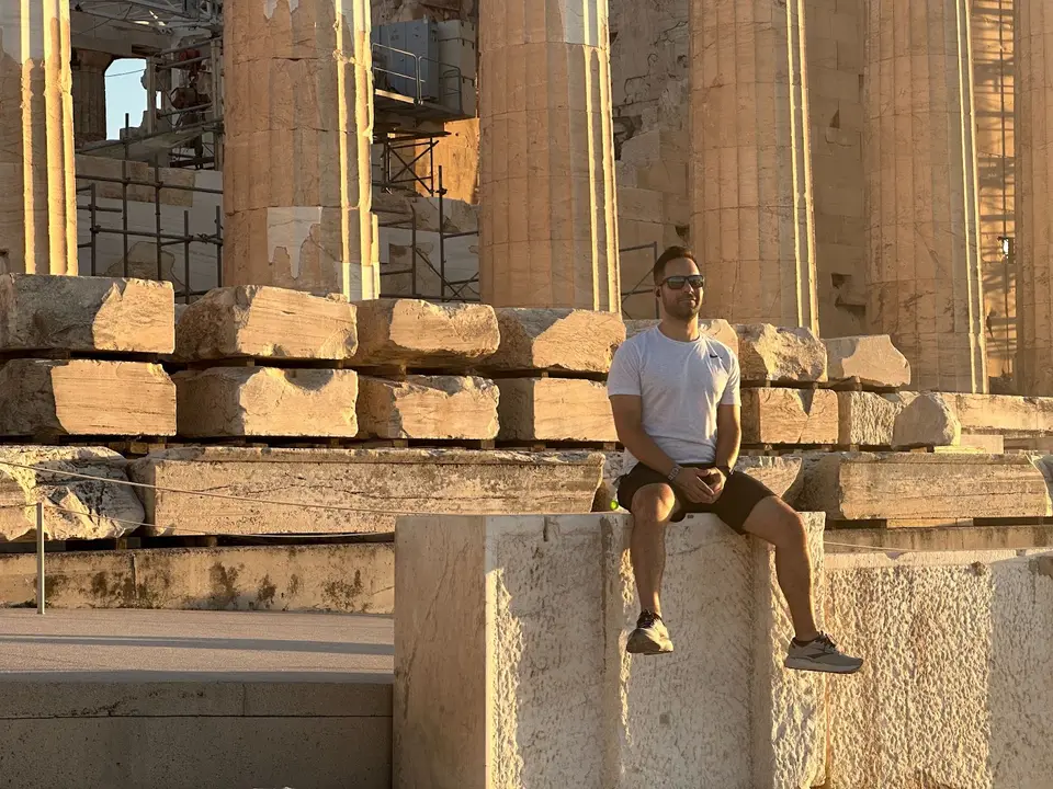 Man in a white t-shirt and sunglasses sitting on ancient stone blocks near tall columns at sunset