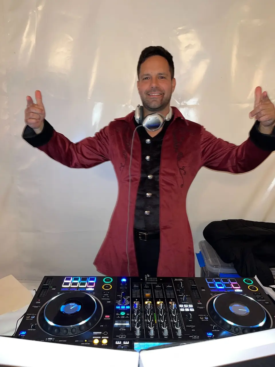 DJ in a burgundy velvet jacket smiling with both thumbs pointing at himself behind a DJ controller