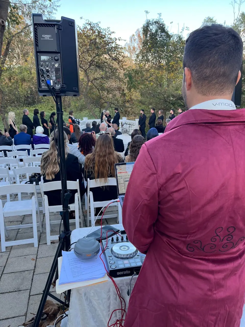 DJ in a burgundy jacket seen from behind, facing seated guests at an outdoor ceremony beside a speaker and controller