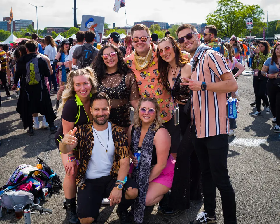 Five friends posing and smiling together at an outdoor festival, one giving a thumbs up