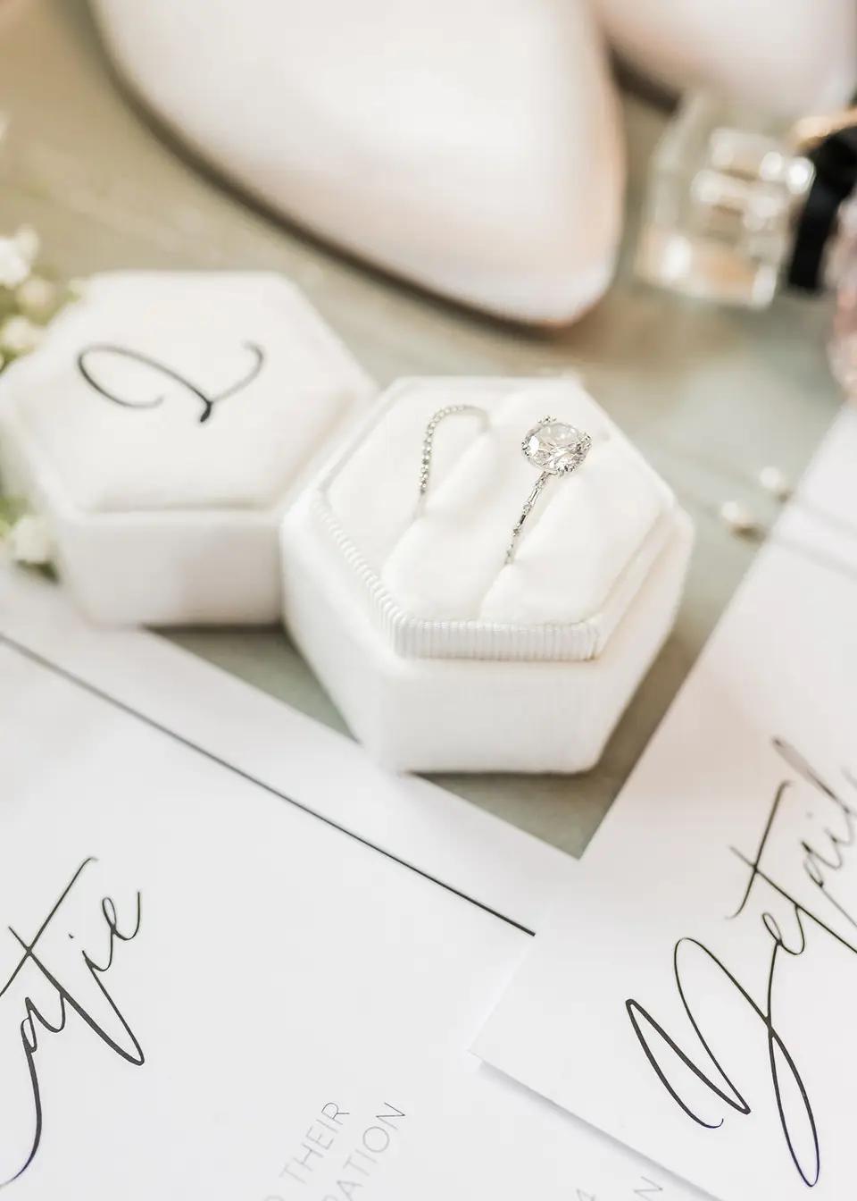 Engagement ring in a white hexagonal ring box on wedding stationery beside bridal shoes