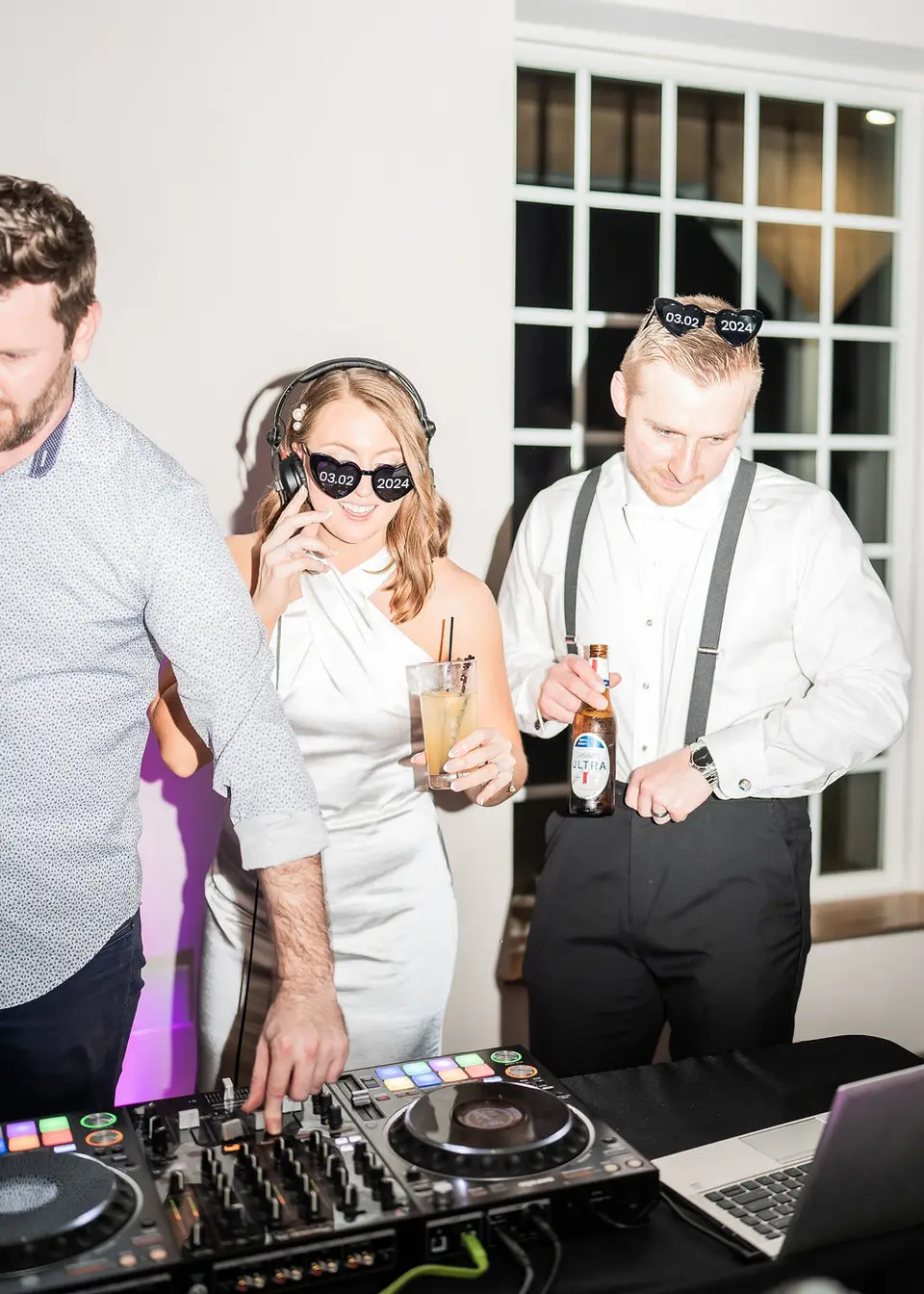 Bride and groom wearing novelty sunglasses at the DJ table, with a man working the controller