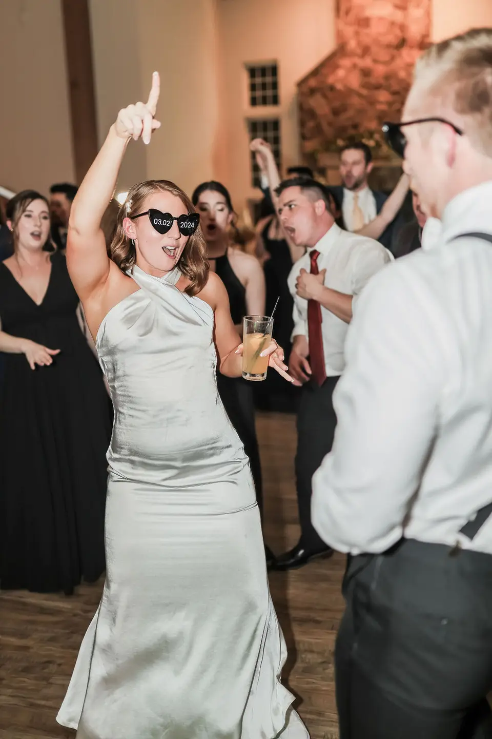 Woman in a satin dress and heart sunglasses points up while dancing, guests behind her