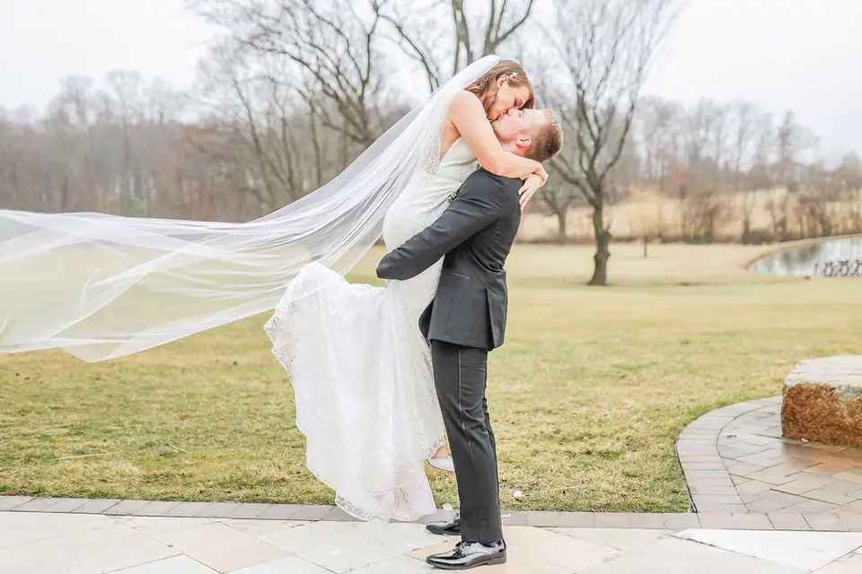 Groom lifts the bride in a lace gown as her long veil blows across a winter field