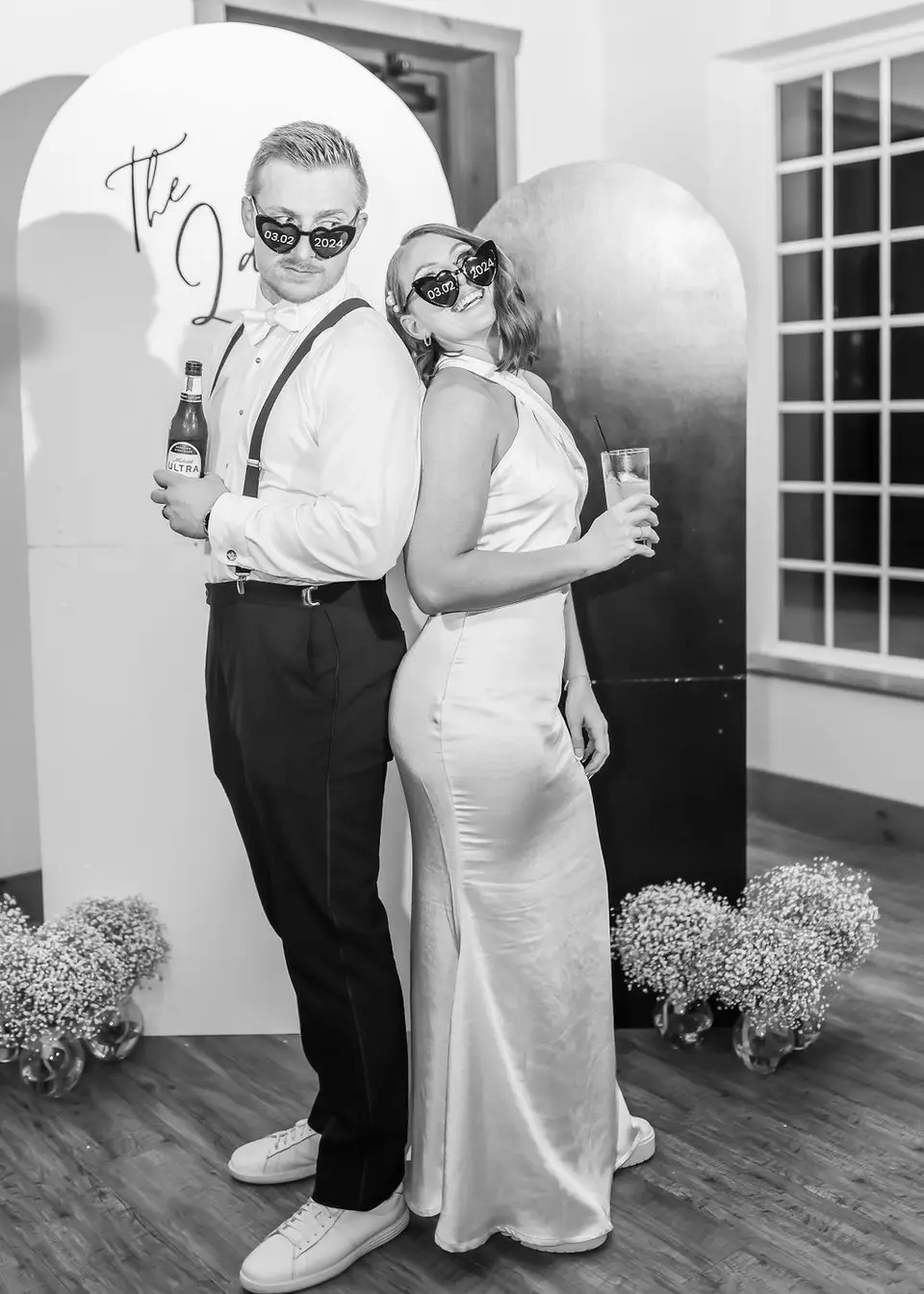 Black and white photo of a couple back to back in novelty sunglasses holding drinks in front of an arch backdrop