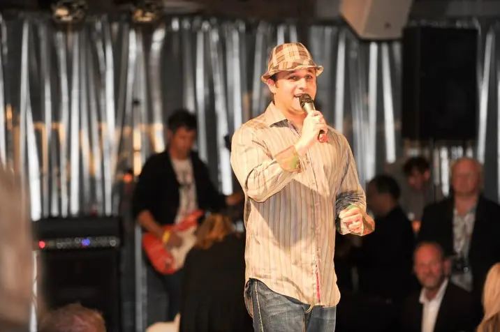 Man in a plaid hat singing into a microphone on stage with a guitarist behind him