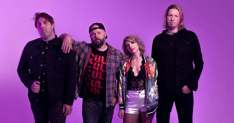 Four band members posing against a purple backdrop, one man in a cap and a woman in a metallic jacket