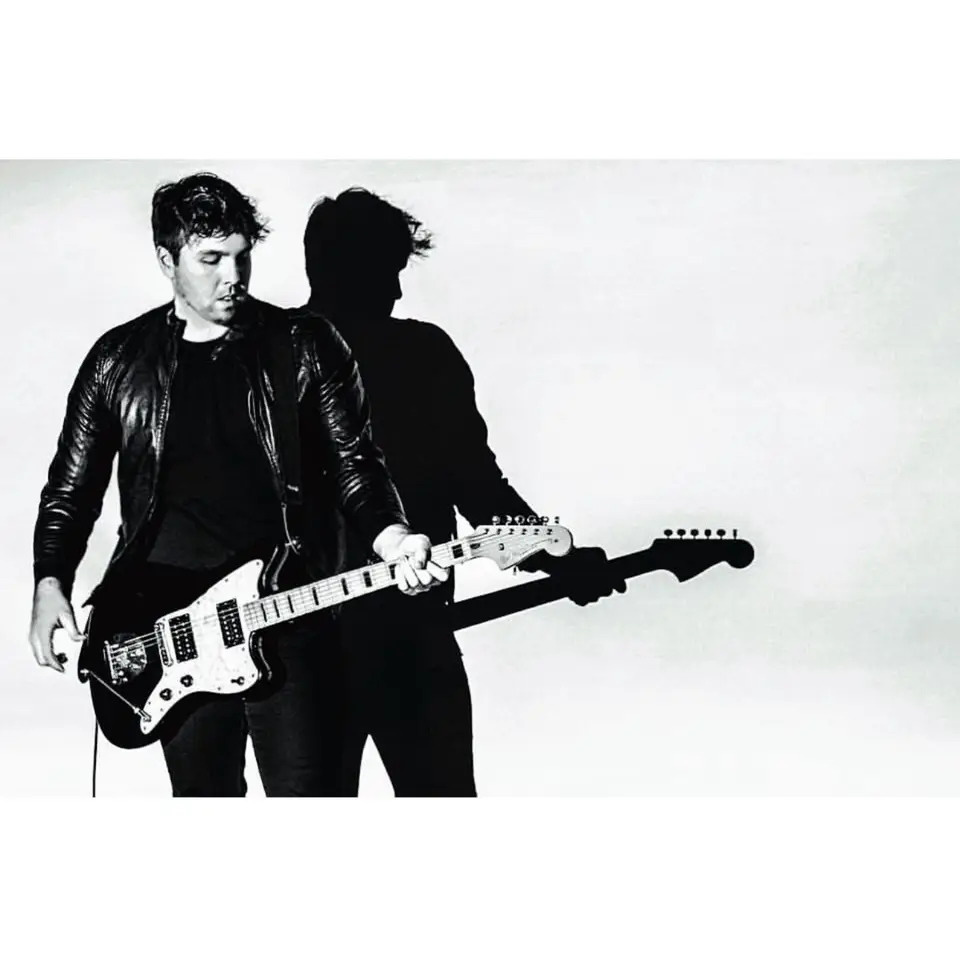 Black and white photo of a man in a leather jacket playing an electric guitar with his shadow behind him