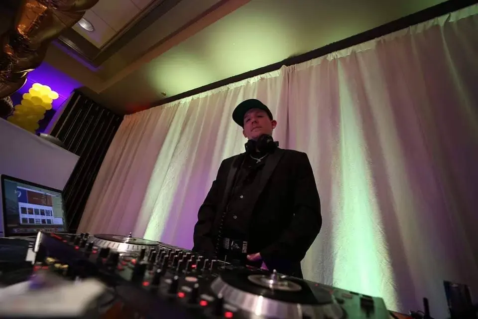 DJ in a black jacket and cap standing behind turntables in front of white draped curtains