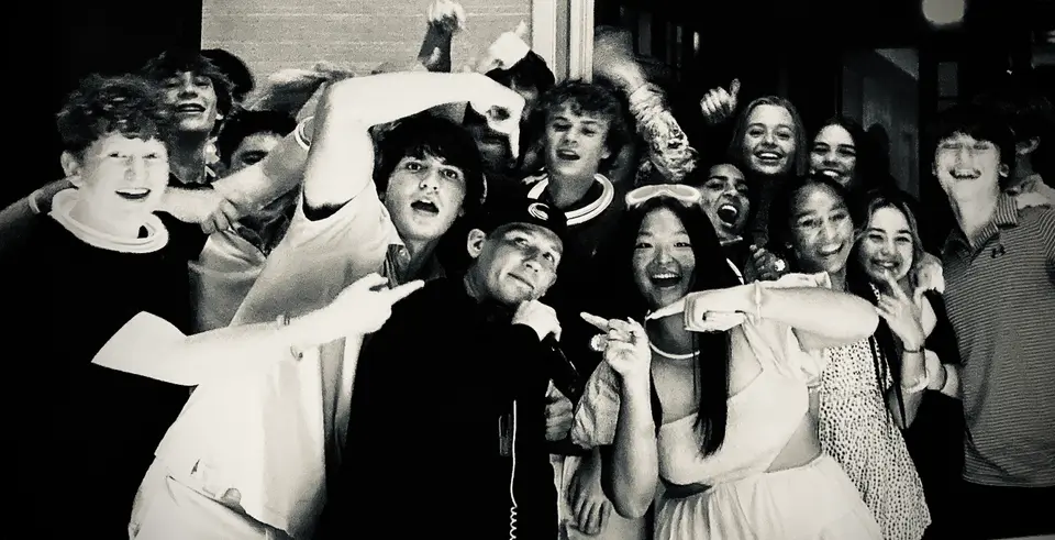 Black and white photo of a DJ in a cap with a microphone surrounded by cheering teenagers