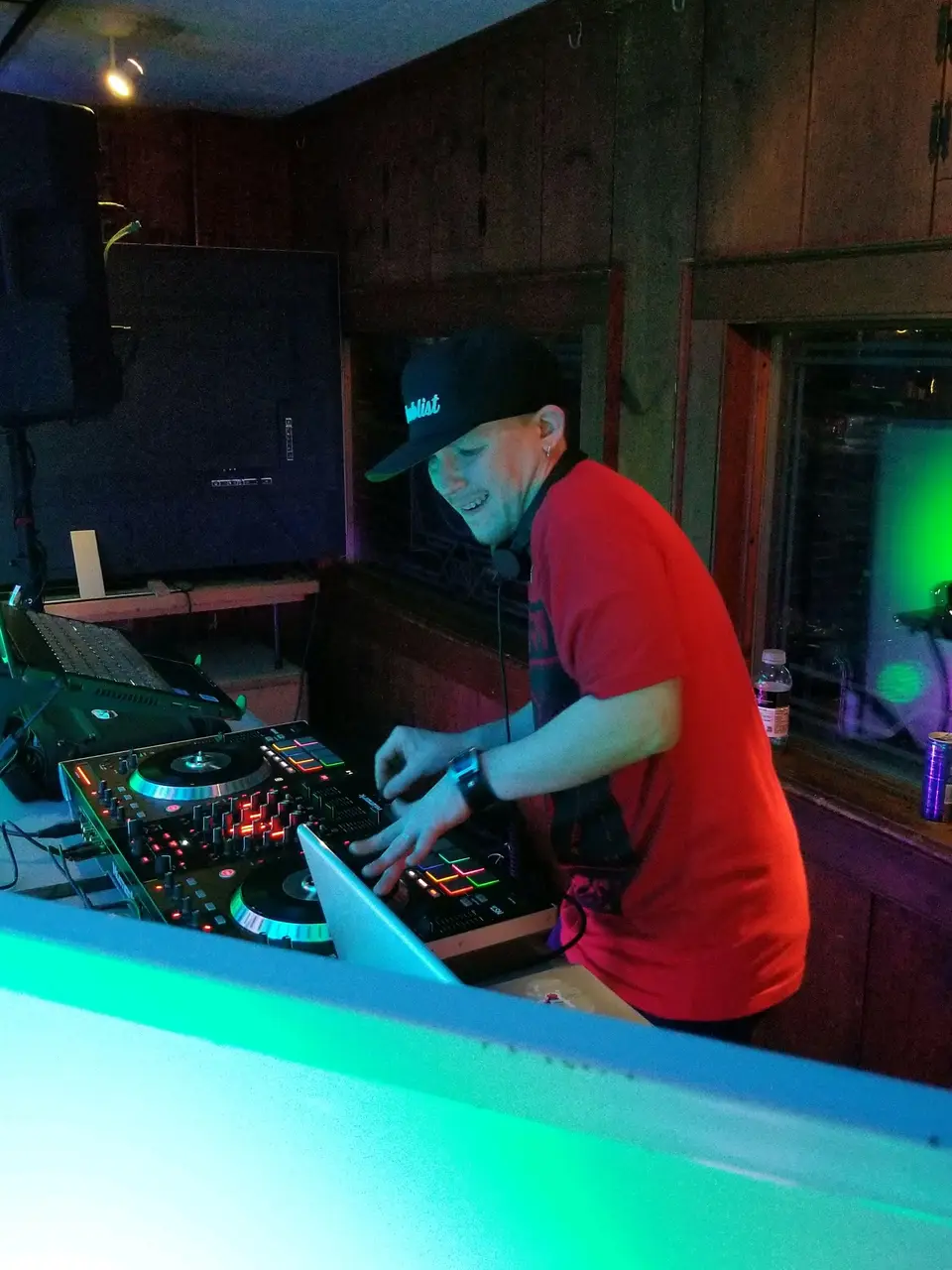 Smiling DJ in a red shirt and cap mixing on a controller in a wood-paneled room with green lighting