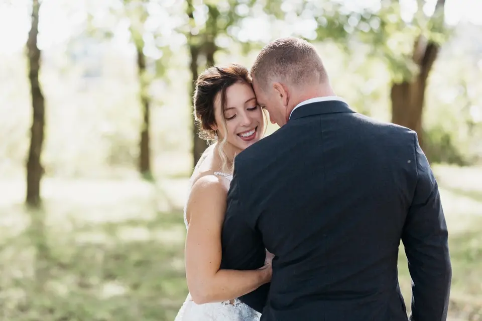 Bride smiling with her arms around the groom in a sunlit grove of trees