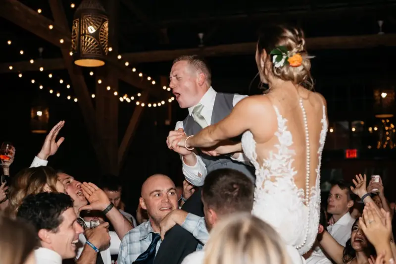 Groom and bride lifted in chairs above cheering guests during a barn wedding reception