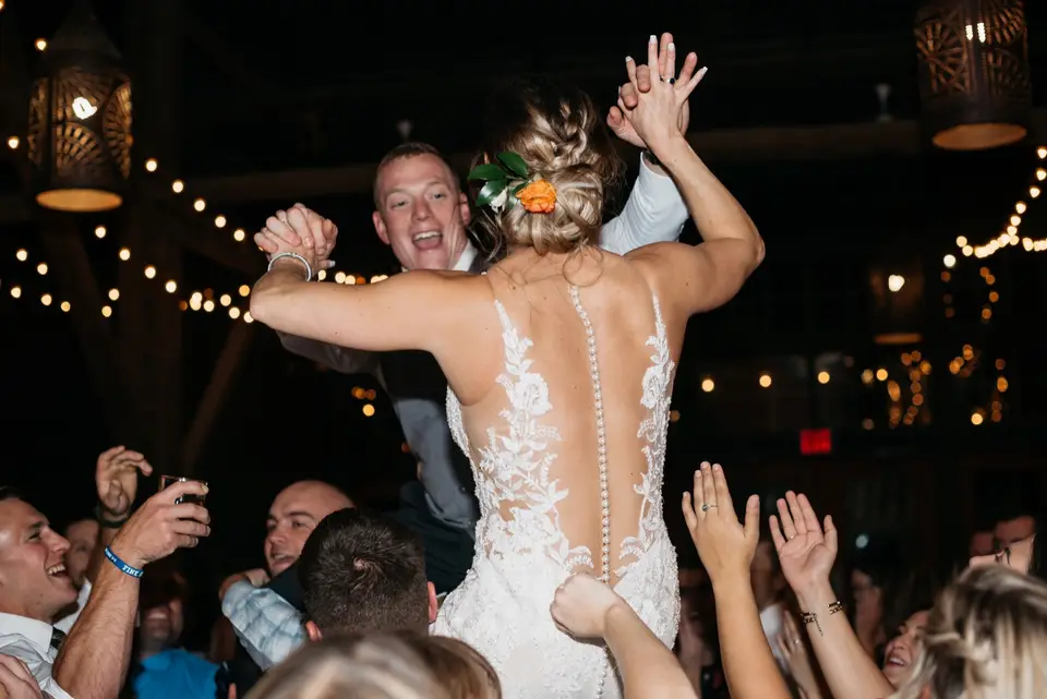 Bride lifted high in the air by guests under string lights in a rustic barn reception