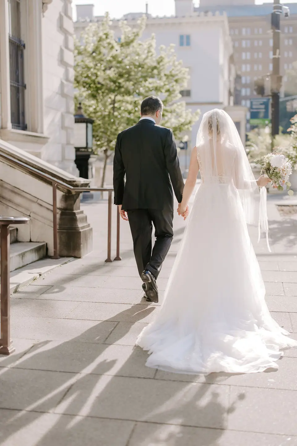 Bride and groom walking away hand in hand on a sunlit city sidewalk, her veil trailing