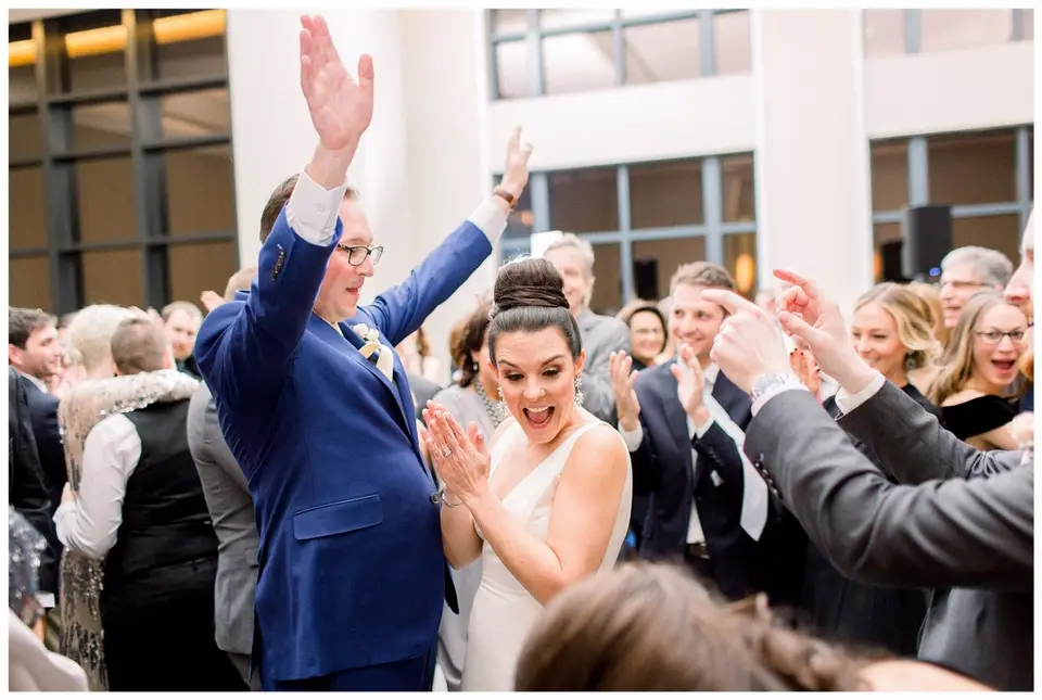 Groom in a blue suit and bride cheer with hands raised while guests surround them on the dance floor