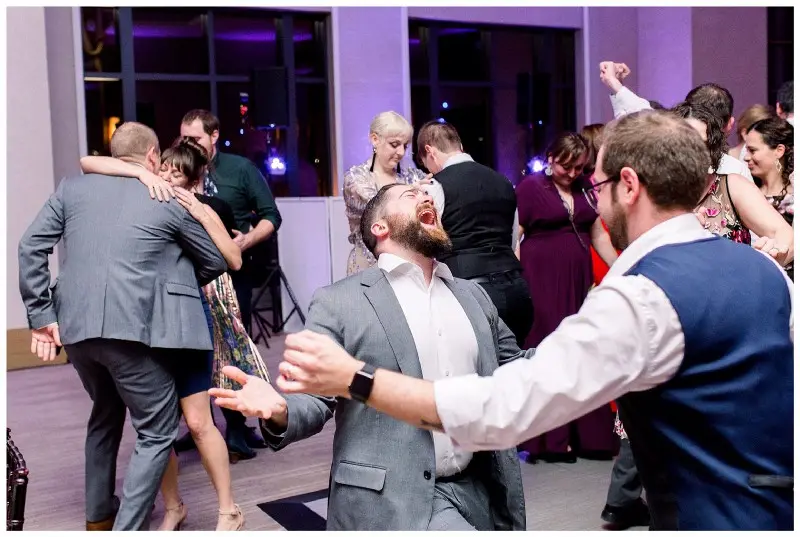 Guests dance with a man in a gray suit mid-shout on a ballroom floor with purple lighting