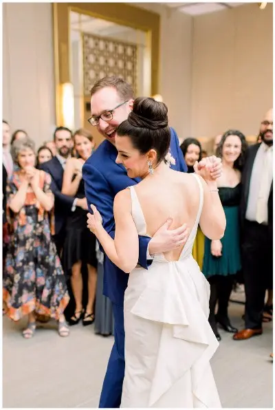 Groom in a blue suit and bride in a white gown share a first dance as guests watch