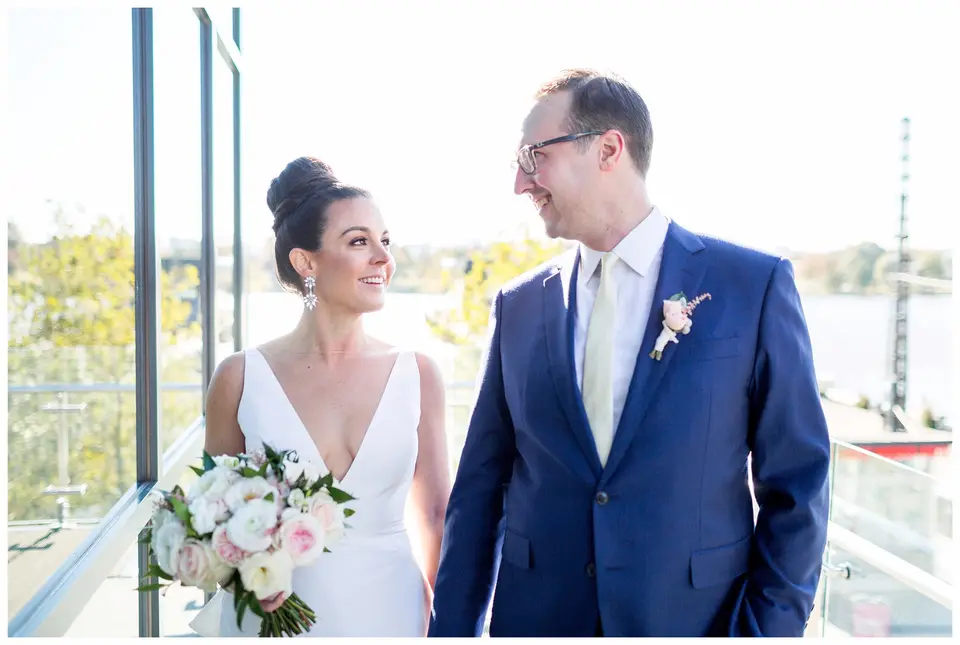 Bride and groom smile at each other on a glass-walled terrace overlooking the water
