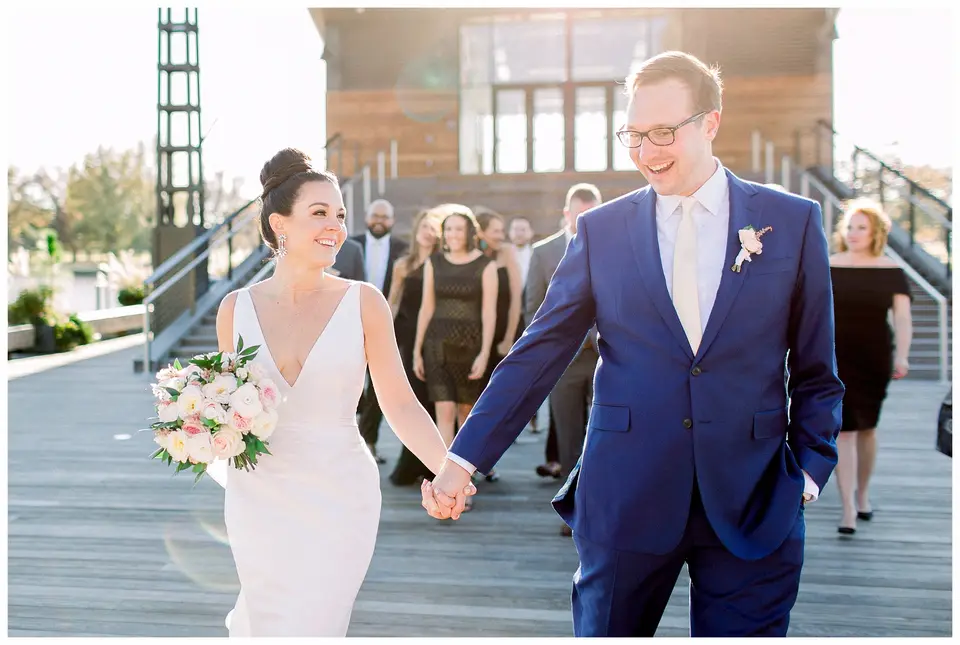 Bride and groom hold hands and smile while walking on a waterfront deck, guests behind them