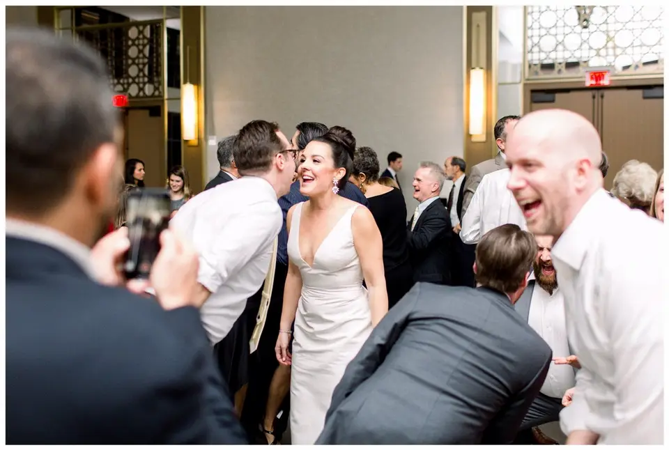 Bride in a white gown laughs while guests dance around her in a ballroom