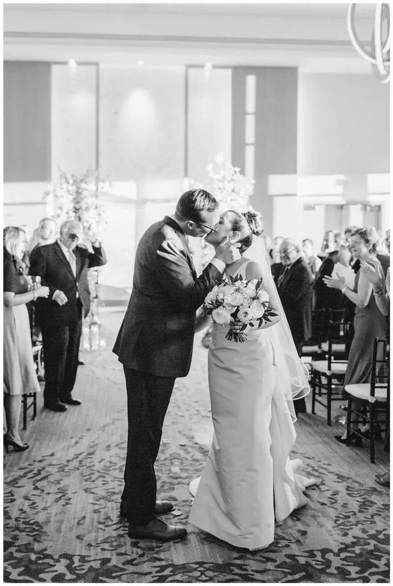 Black and white photo of the bride and groom kissing in the aisle as guests applaud