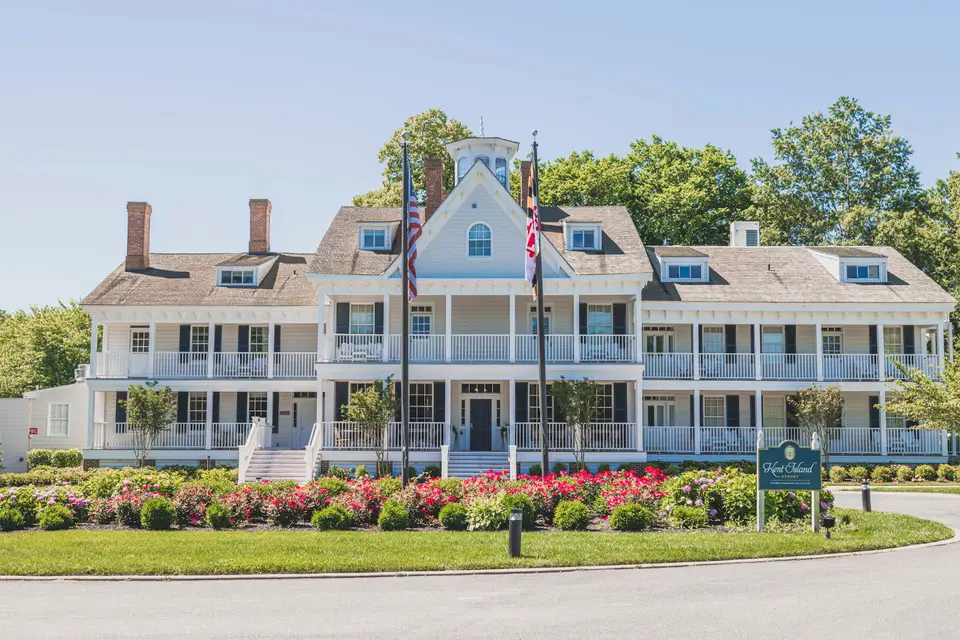 White two-story venue with wraparound porches, flagpoles and flower beds under a clear blue sky