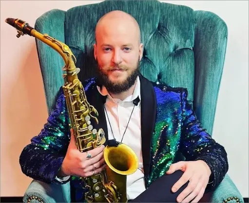 Bearded saxophonist in a sequin jacket seated in a teal armchair holding an alto saxophone