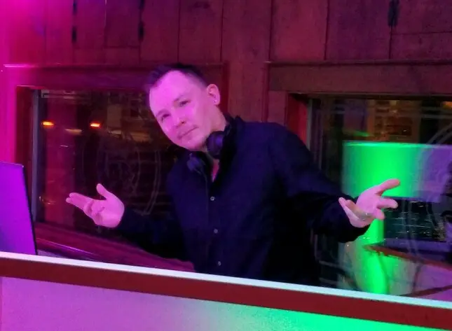 DJ in a dark shirt with headphones around his neck, shrugging with arms out behind a purple-lit booth