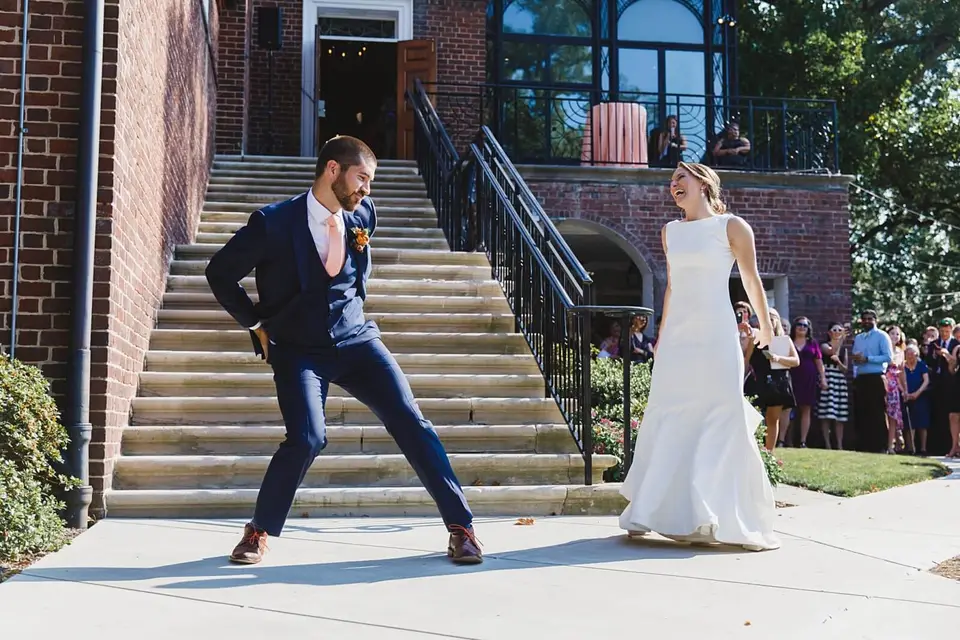 Groom in a navy suit dancing playfully on a sidewalk as the bride in a white gown laughs