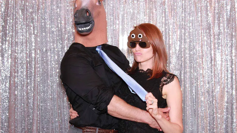 Man in a horse mask and woman in poop emoji sunglasses posing at a photo booth with a sparkly backdrop