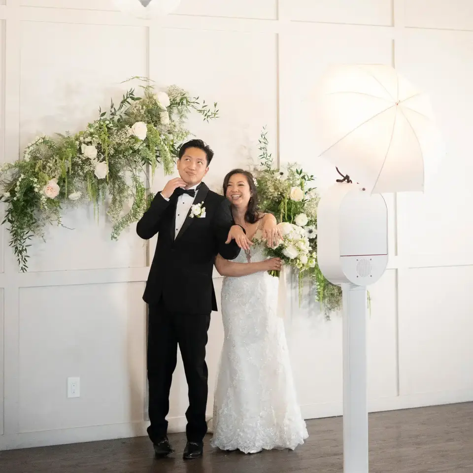 Bride and groom standing beside a white photo booth with a floral backdrop and umbrella light